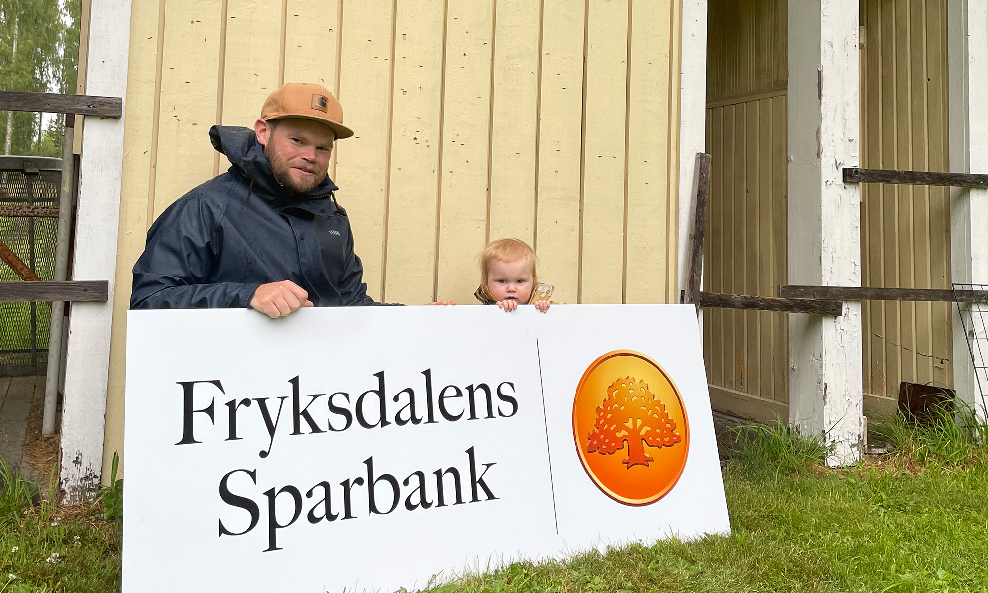 En man och ett barn håller en vit skylt med Fryksdalens Sparbanks logo på.